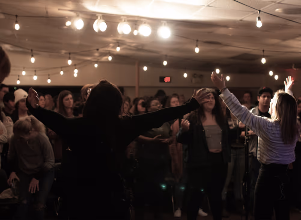 People gathered indoors at an event with string lights overhead, some raising their hands in the air.