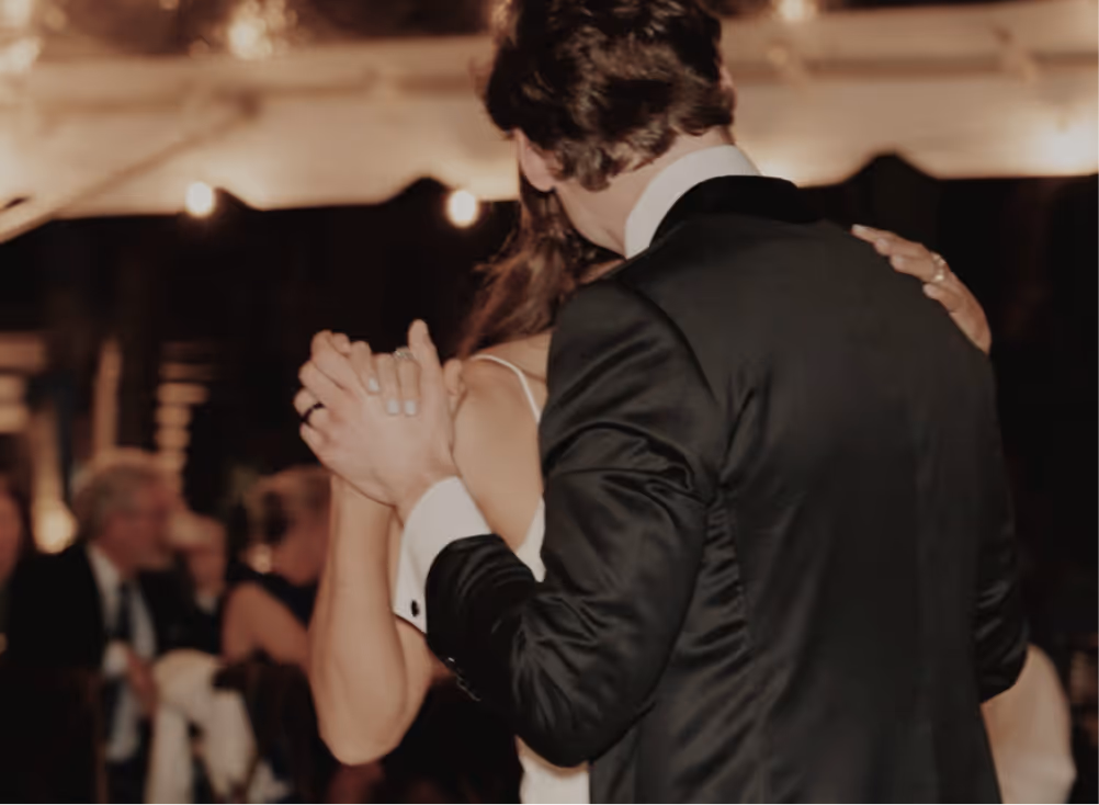 Bride and groom in formal attire sharing a slow dance at a wedding reception with blurred guests in the background.