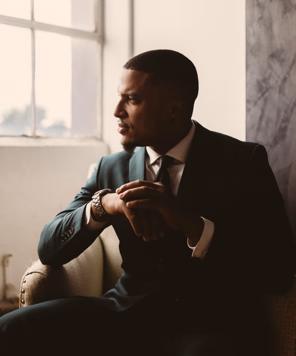 Man in a dark suit sitting on a chair, looking pensively out a window with natural light.