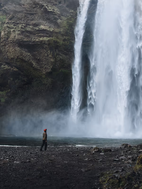 Individual wearing a red hat stands before a majestic waterfall tumbling down a rocky cliff amidst misty surroundings