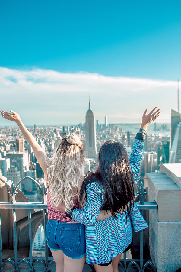 Two women standing on a rooftop observation deck in New York City with arms raised, celebrating the breathtaking skyline