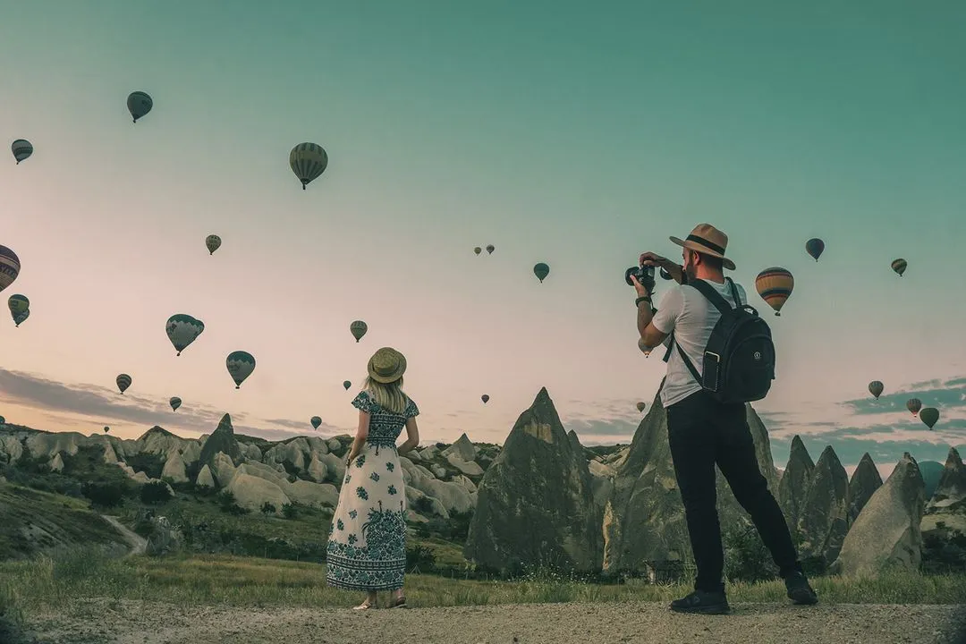 Couple standing among stunning rock formations with colorful hot air balloons filling the sky during sunrise or sunset