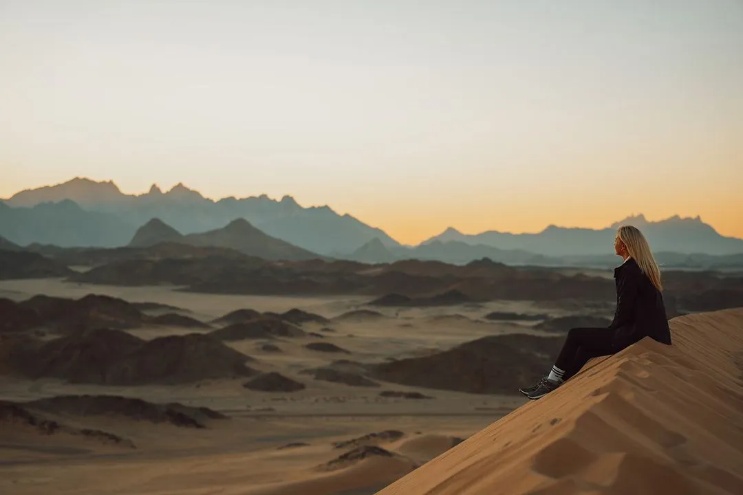 A lone traveler sits atop a sand dune at sunset, overlooking a sprawling desert landscape with rugged mountains in the distance