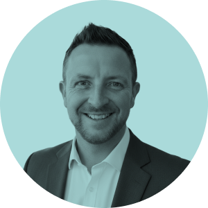 Smiling man with short hair wearing a dark suit jacket and white shirt against a light blue background.