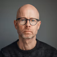 Portrait of a middle-aged bald man with glasses and a trimmed beard wearing a dark sweater against a neutral background.