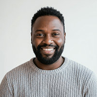 Smiling Black man with short curly hair and beard wearing a gray knit sweater against a light background.