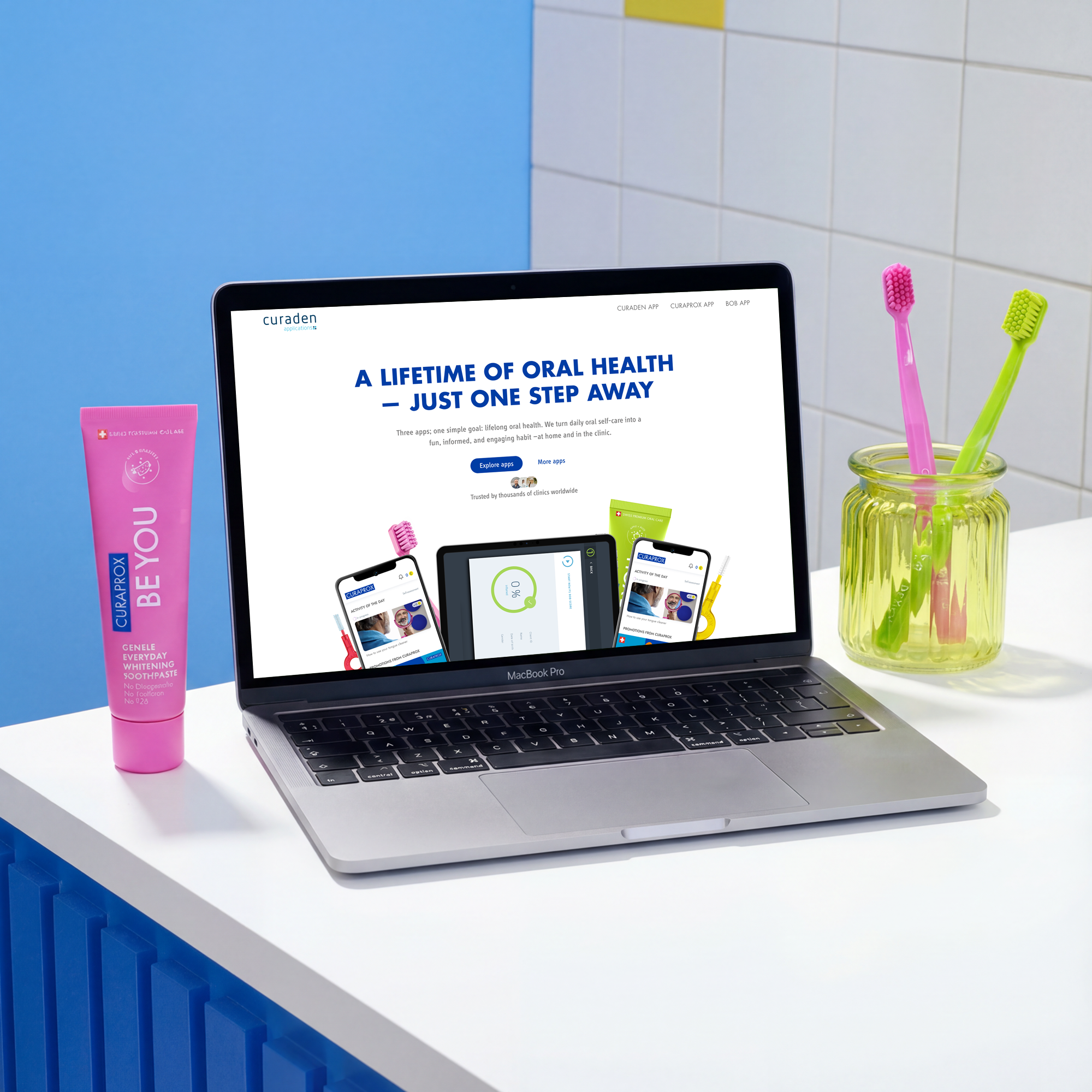 Open MacBook Pro on a white table displaying a dental health website, with a tube of pink Curaprox BE YOU toothpaste on the left and a jar holding two colorful toothbrushes on the right.