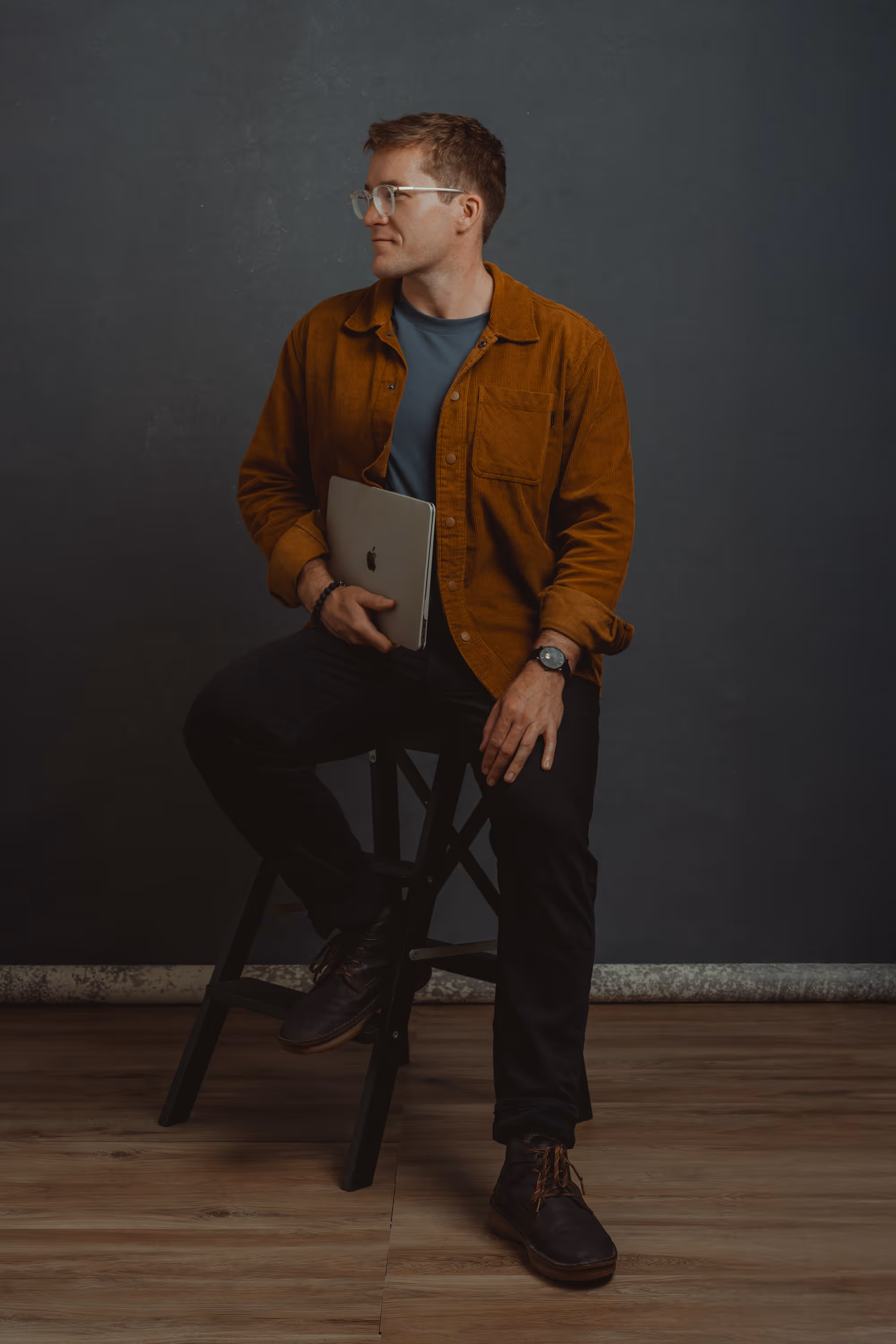 Young man wearing glasses, brown jacket, and black pants sitting on a wooden stool holding a closed silver MacBook.