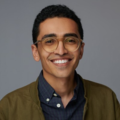 Smiling young man with short curly hair and glasses wearing a dark blue shirt and olive green jacket against a gray background.