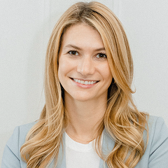 Smiling woman with long blonde hair wearing a white top and light blue blazer against a plain background.