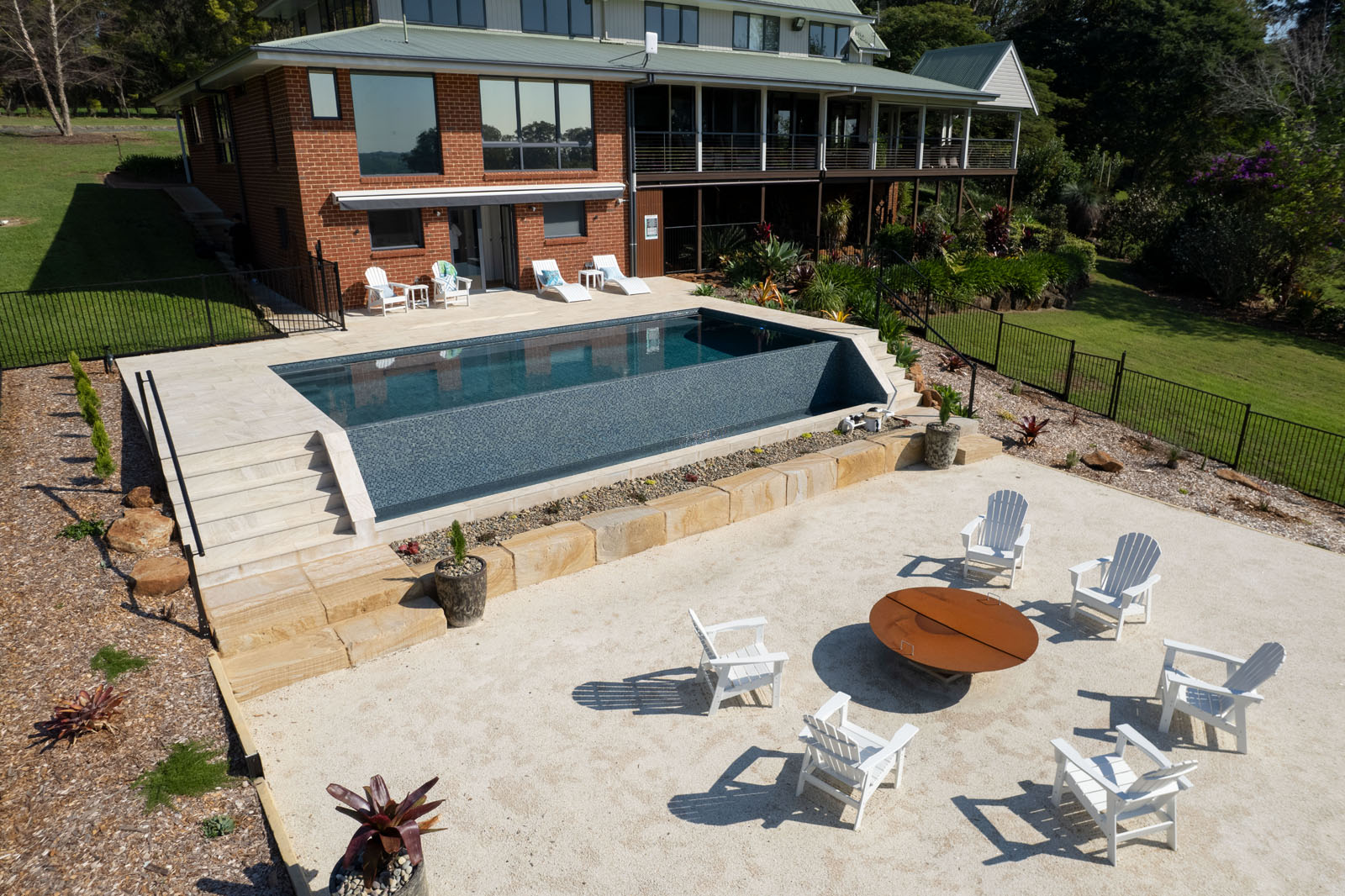 An aerial view of a large house and  serene pool overlooking a lush green field under a clear blue sky.