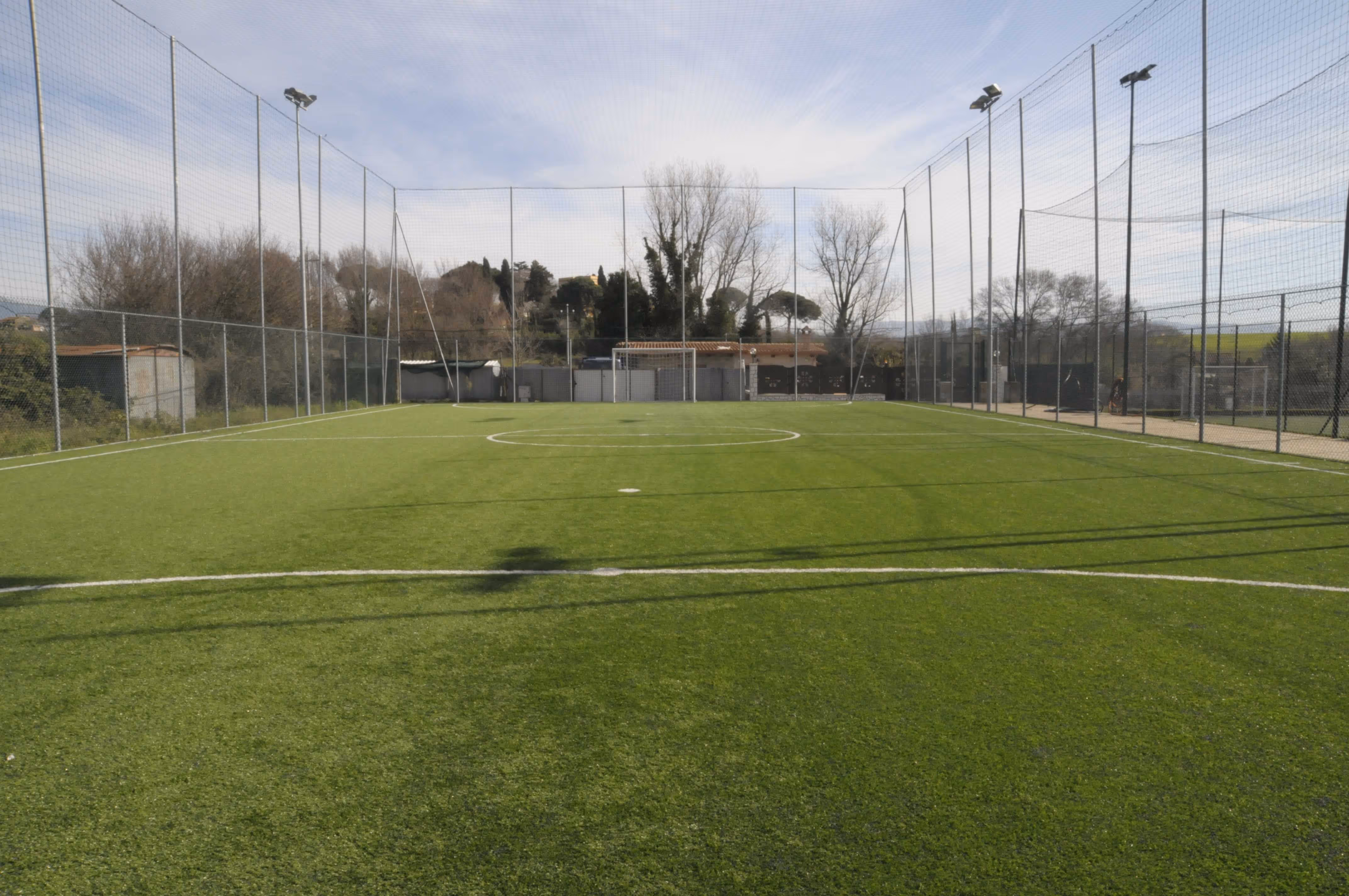 Campo da calcio all'aperto con rete di protezione e cielo parzialmente nuvoloso.