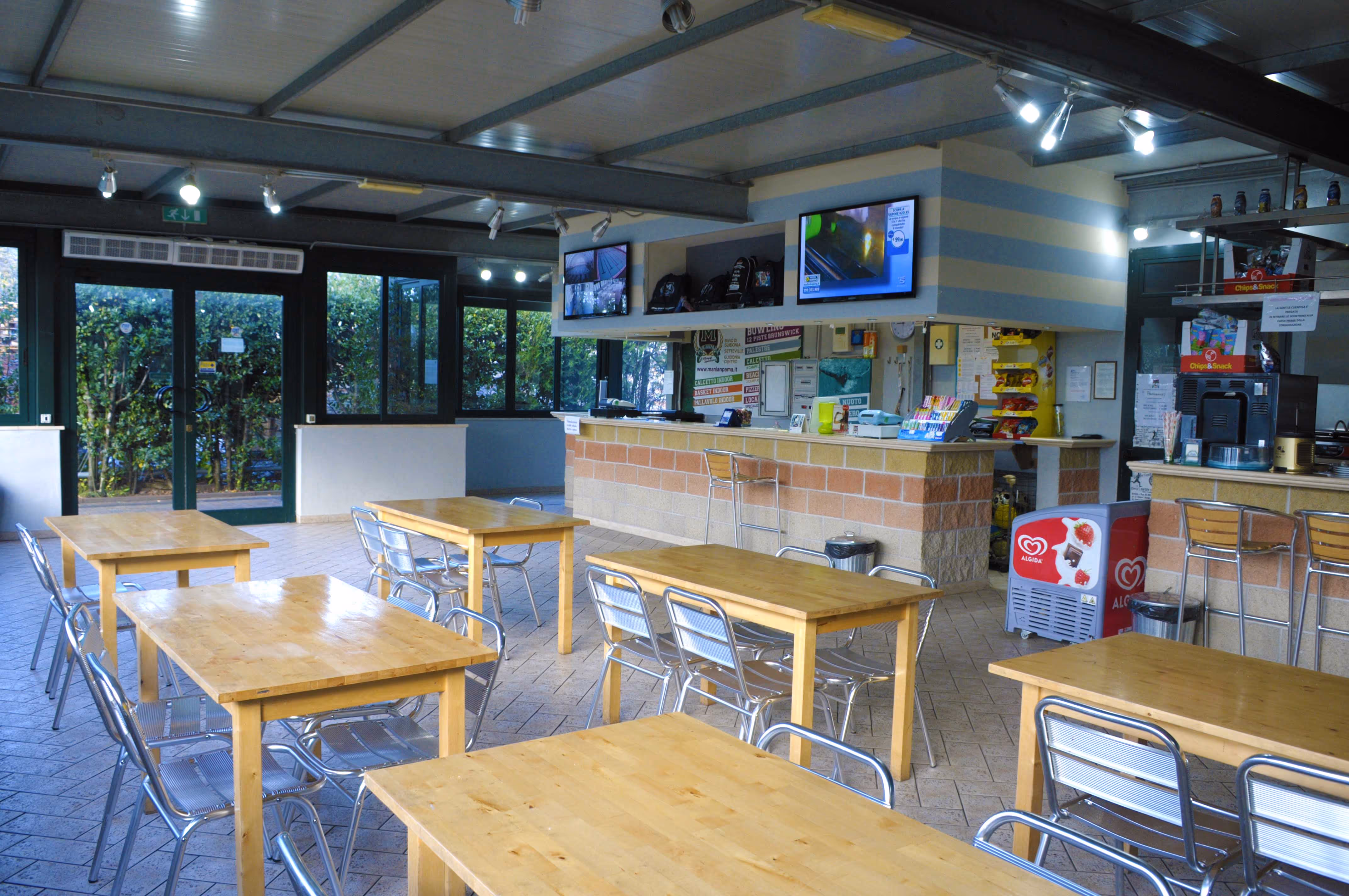 Sala interna di una caffetteria con tavoli di legno e sedie metalliche, bancone con monitor sopra e un frigorifero per gelati Algida.