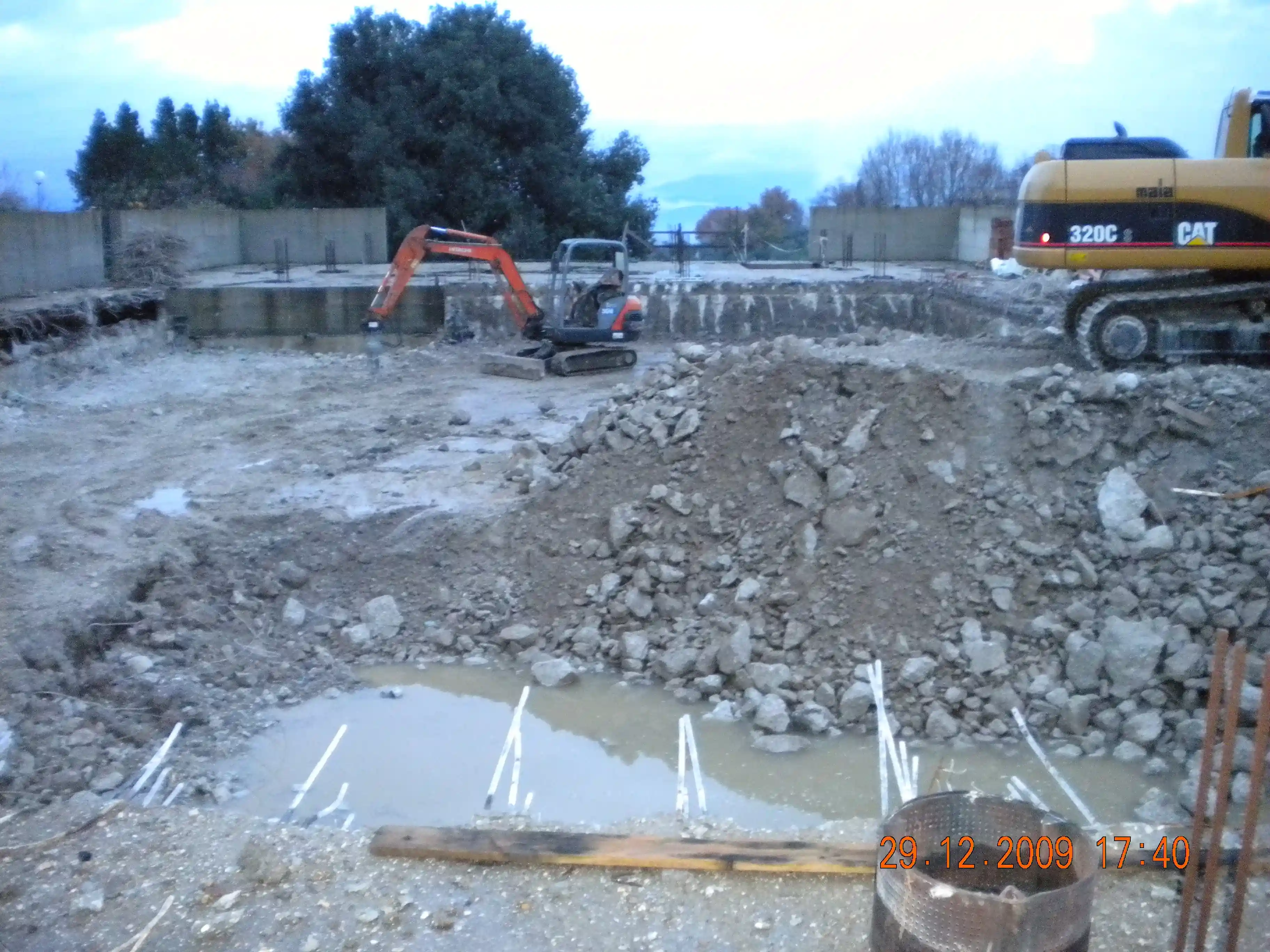 Cantiere edile con escavatori, cumulo di terra e pozzanghere d'acqua sotto cielo nuvoloso.
