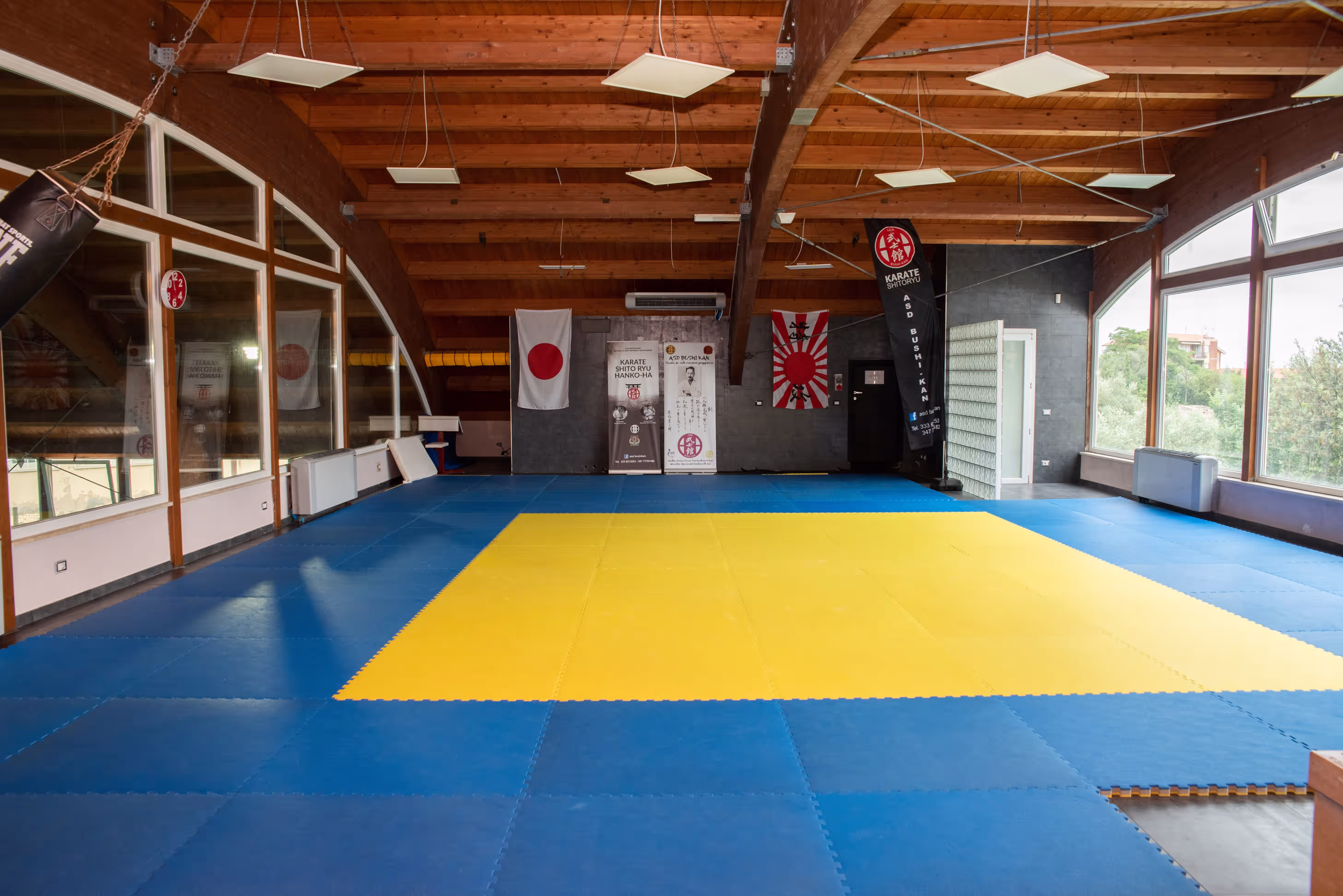 Sala di allenamento di karate con pavimento imbottito blu e giallo, bandiere giapponesi appese e pareti con grandi finestre.