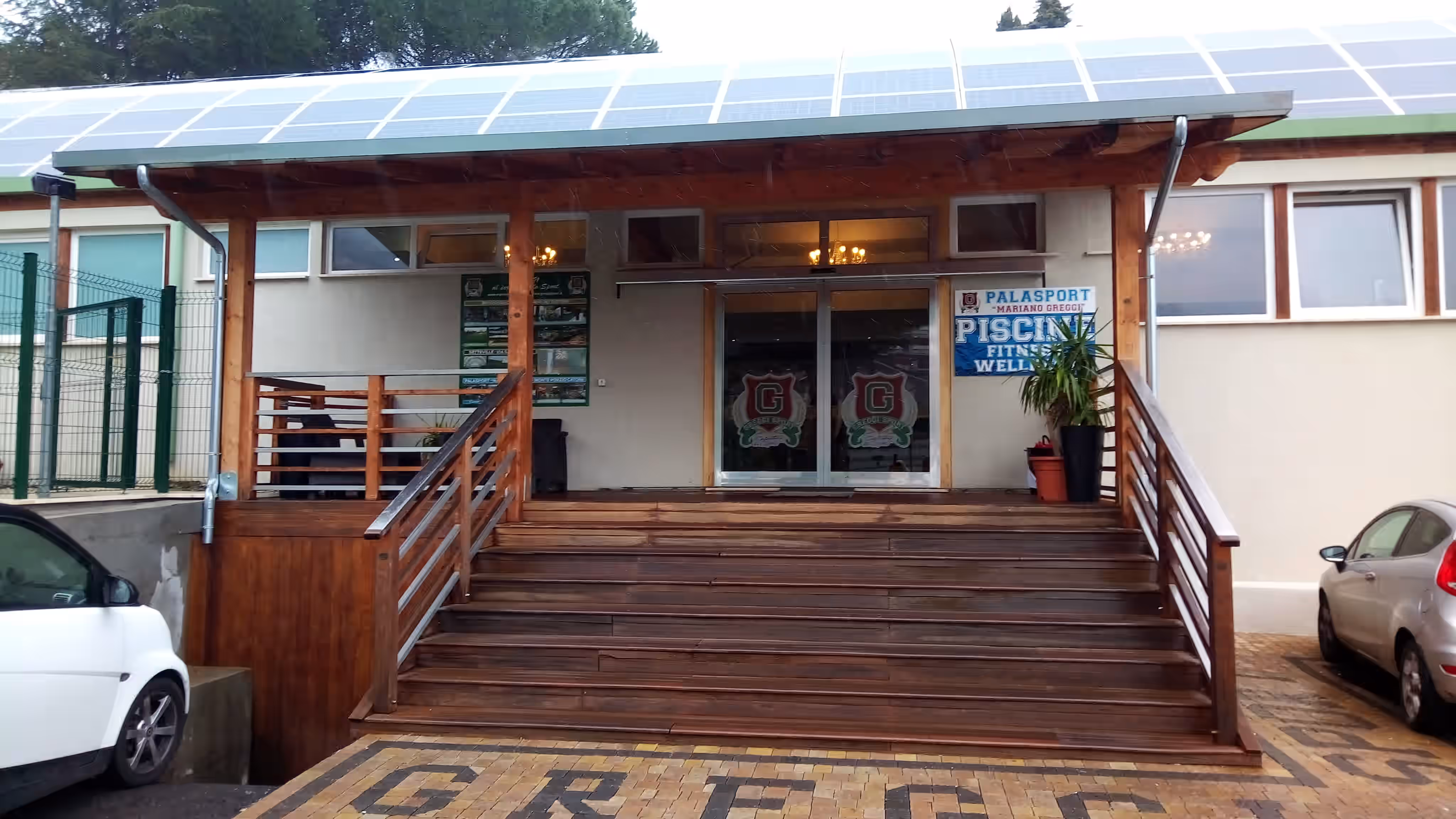 Ingresso di un edificio con ampie scale in legno, tetto con pannelli solari e due auto parcheggiate ai lati.