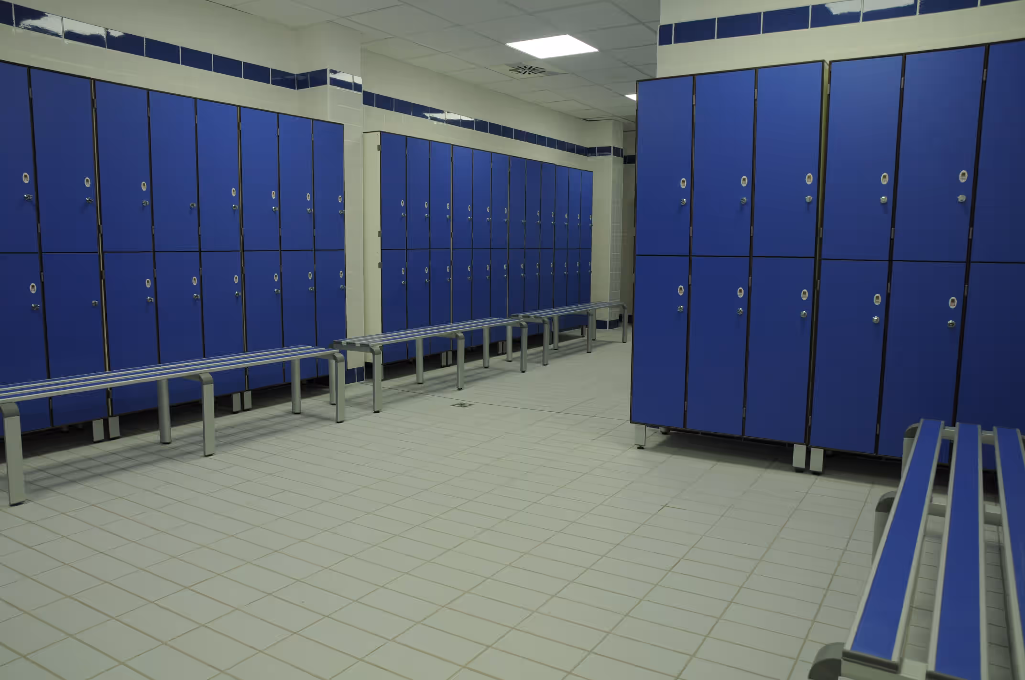 Spogliatoio con armadietti blu e panchine metalliche con sedili blu su pavimento piastrellato chiaro.