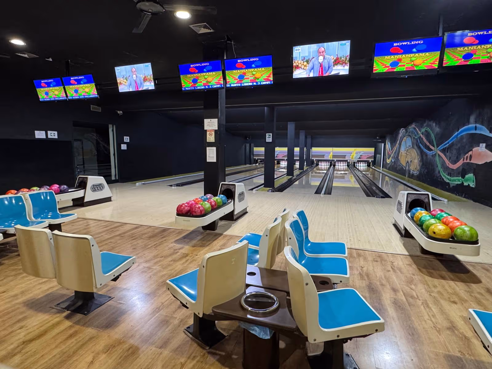 Interno di una pista da bowling vuota con sedili blu, palle da bowling colorate e monitor sopra le piste.