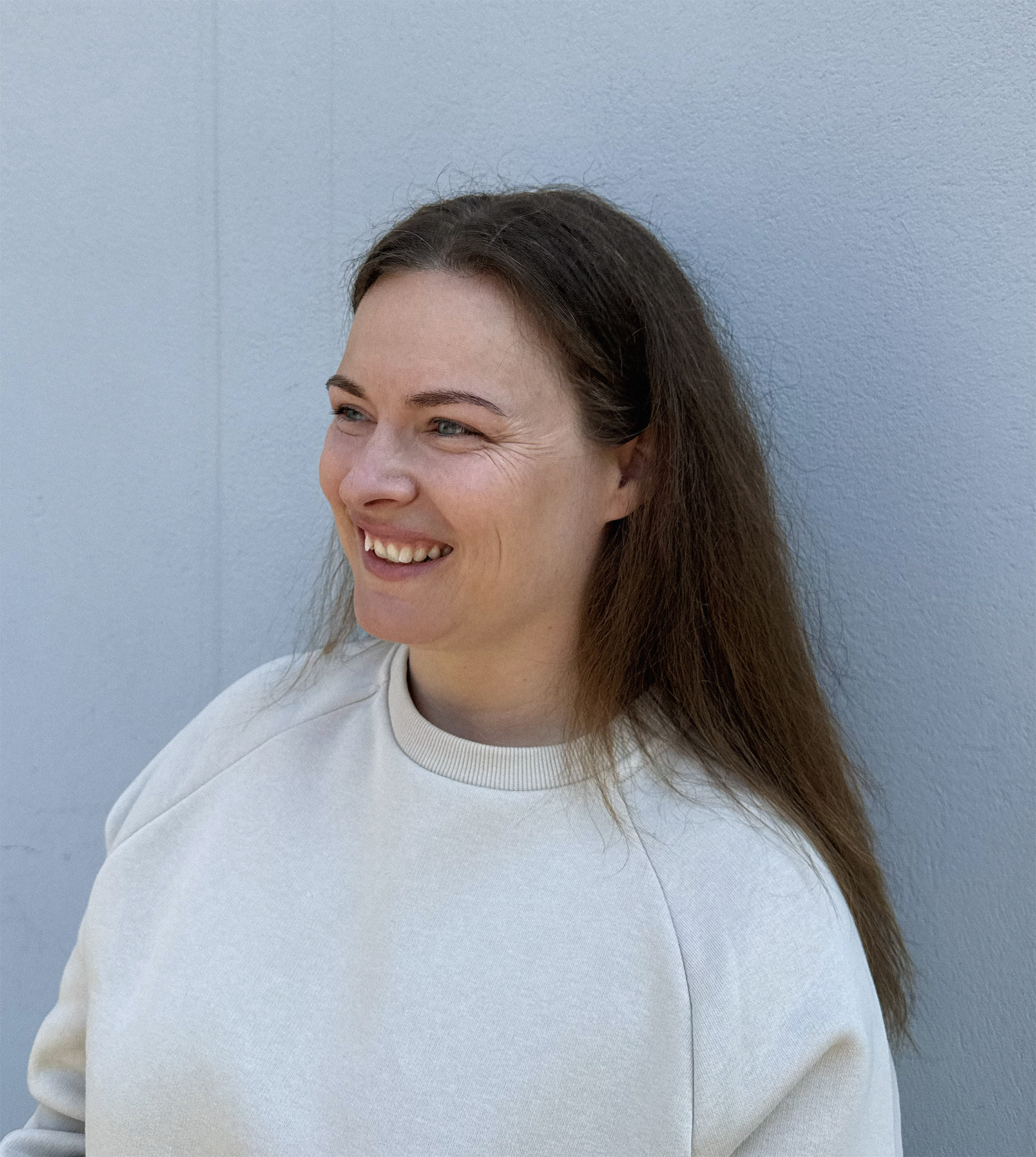 Woman with long brown hair smiling and looking to the left, wearing a light beige sweatshirt, in front of a light blue textured wall.