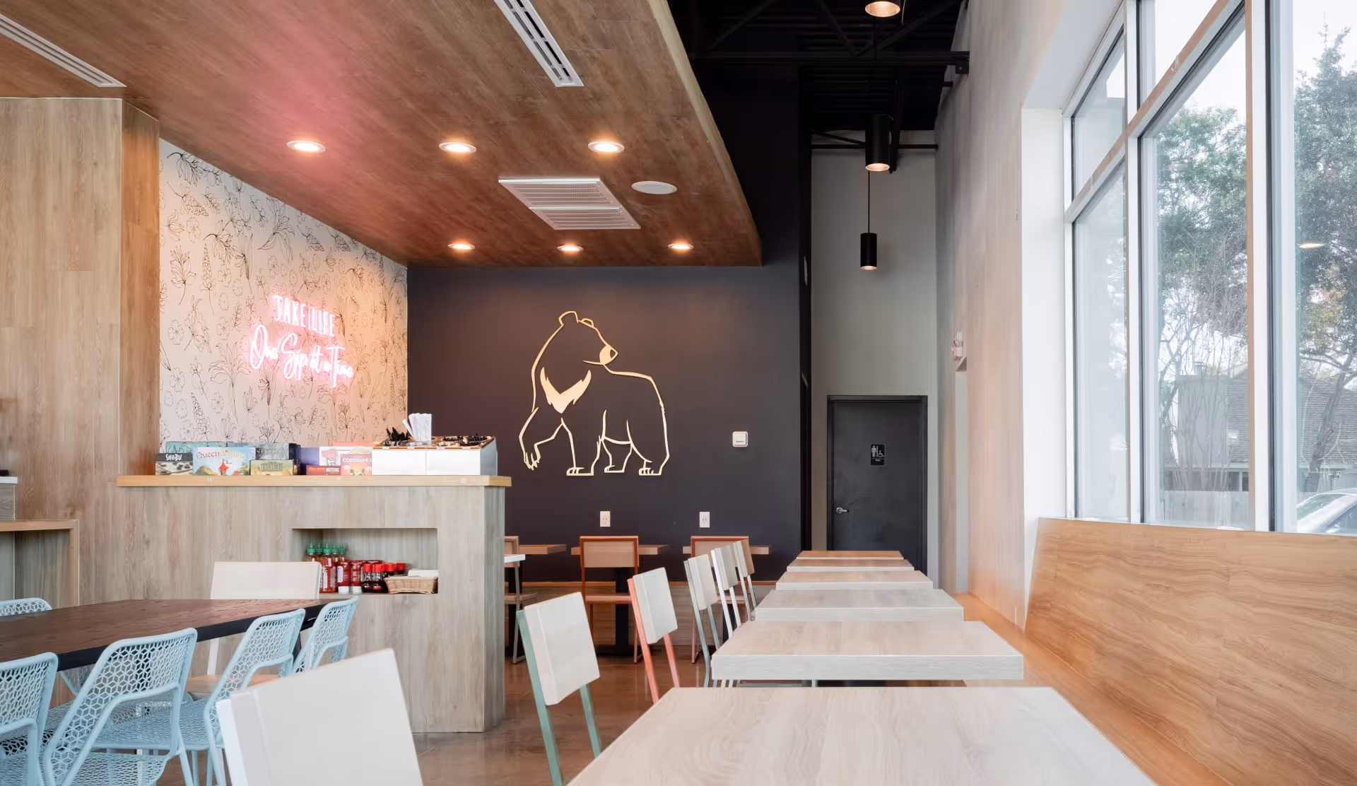 Artea interior with light wooden tables and chairs, a neon sign on a floral wall, and a bear illustration on a dark accent wall.