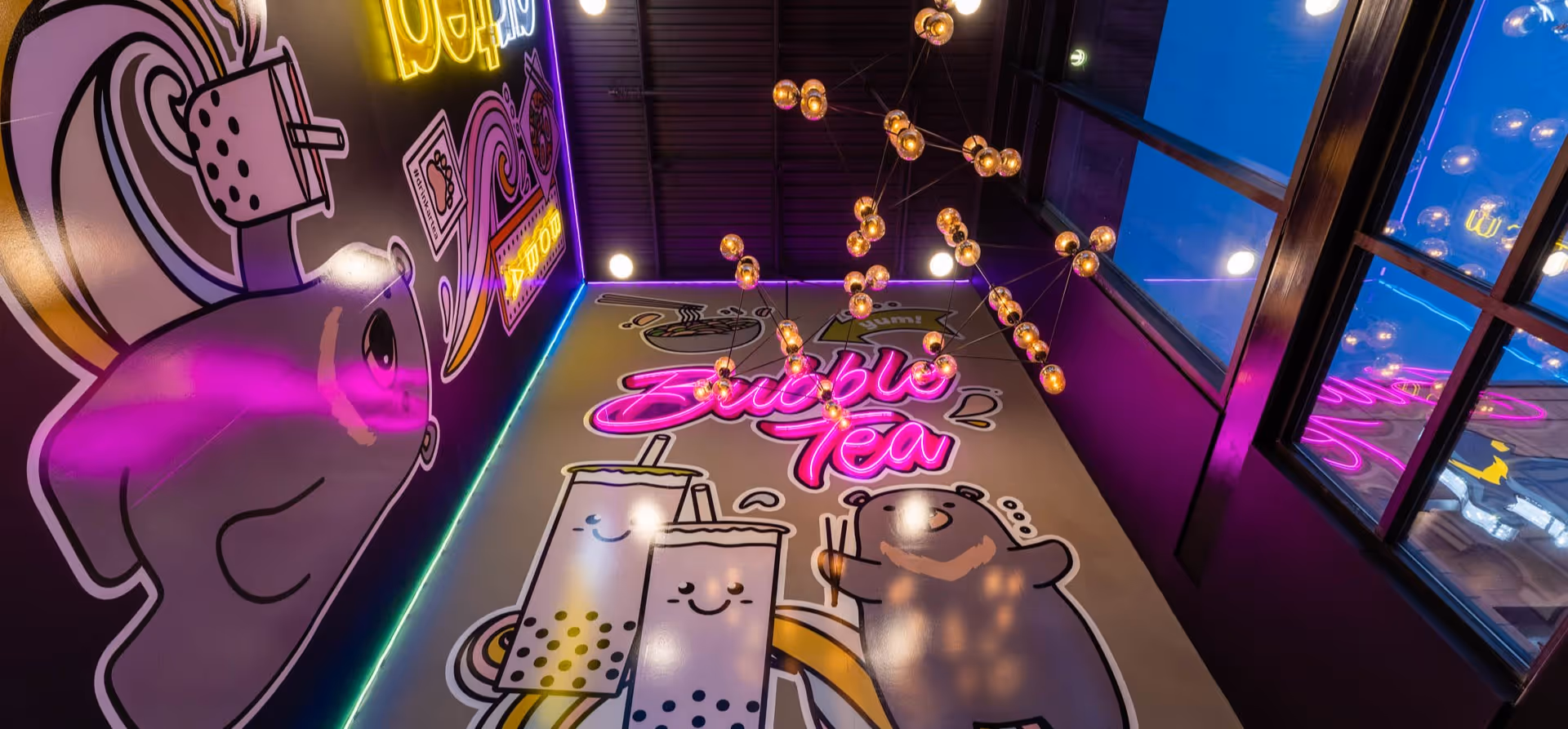 Wall mural with neon pink 'Bubble Tea' text, smiling bubble tea cups, a cartoon bear, and hanging spherical lights.