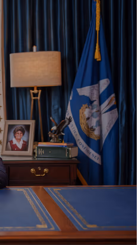 Desk with blue leather writing surface in front of blue curtains and a Louisiana state flag, with a table lamp, eagle statue, books, and a framed photo on a wooden side table.