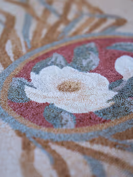Close-up of a floral embroidery with a white flower and green leaves on a beige fabric background.