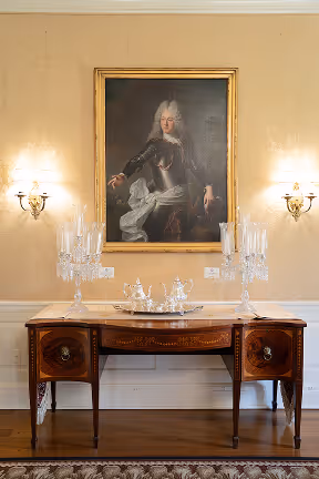 Antique wooden sideboard with crystal candle holders and silverware, beneath a framed portrait of a man in 18th-century attire.