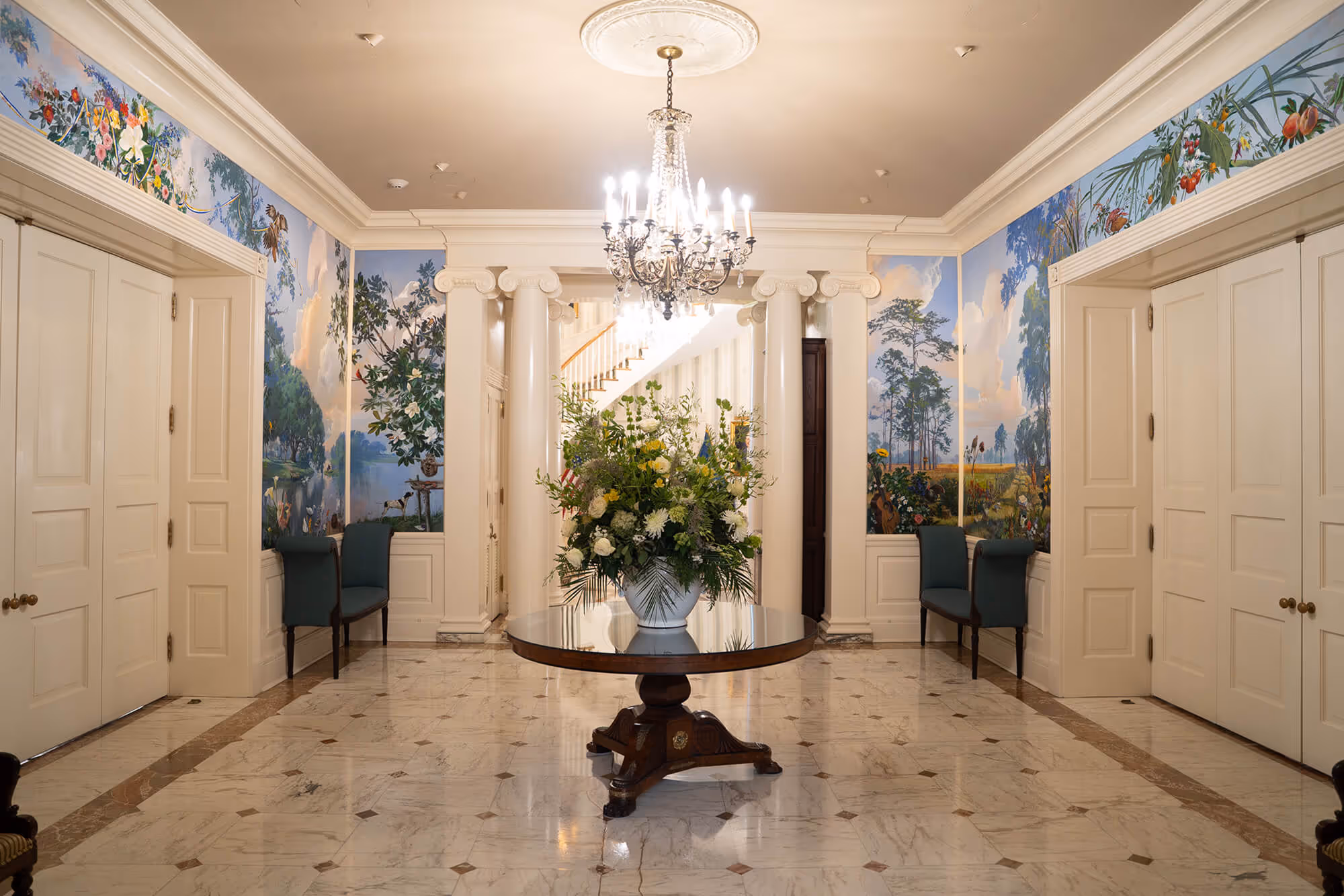 Elegant foyer with marble floor, round wooden table holding a large floral arrangement, white columns, chandelier, and walls adorned with nature-themed murals.