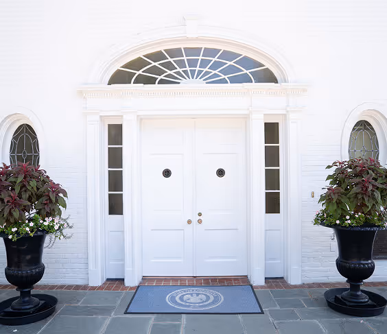 White double front doors with sidelights and a fanlight window above, flanked by two large black planters with green and red foliage.