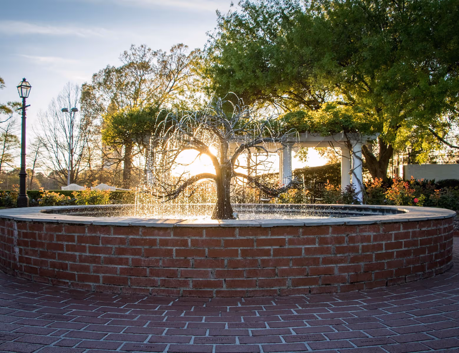 Circular brick fountain with a metal tree sculpture spraying water, set in a garden with greenery and a lamppost.