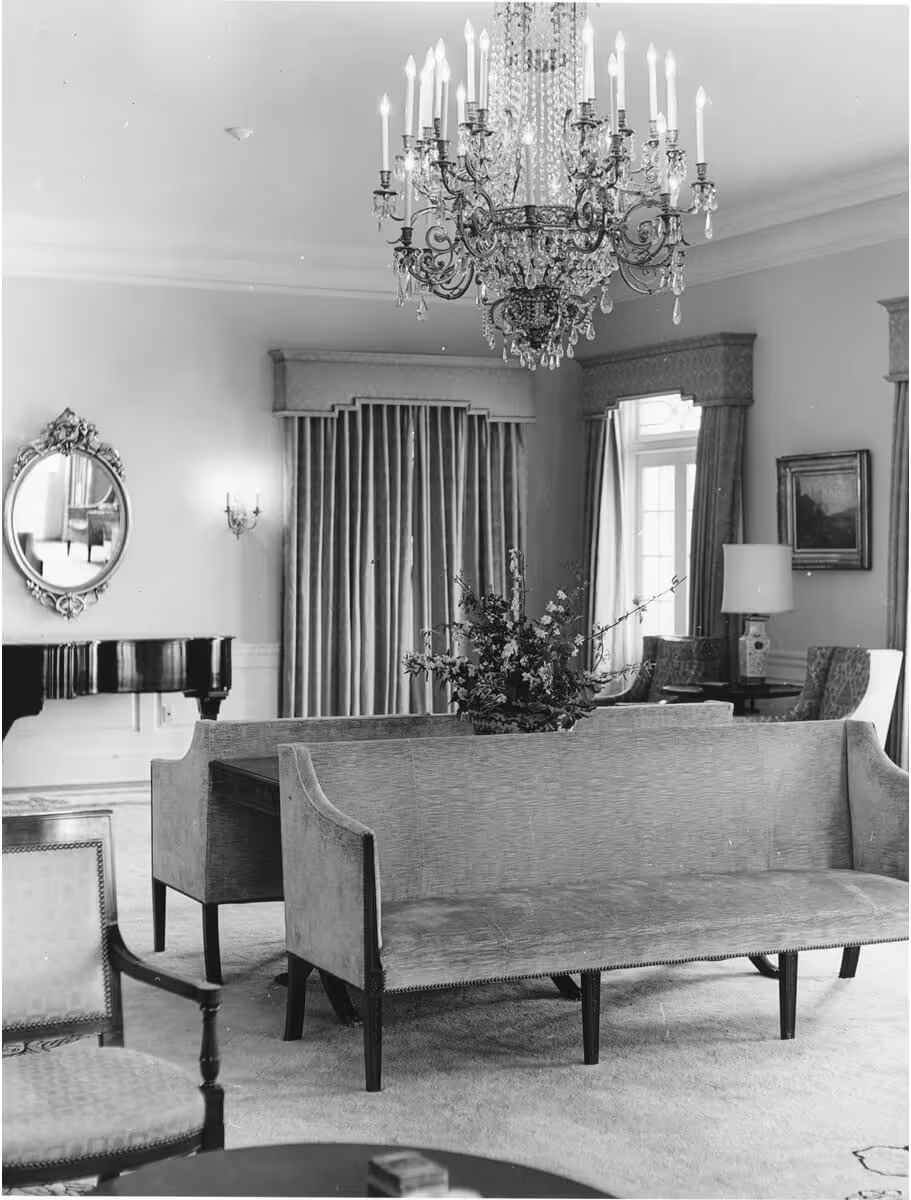 Black and white photo of a formal living room featuring a large chandelier, upholstered couches, a piano, and heavy drapes.