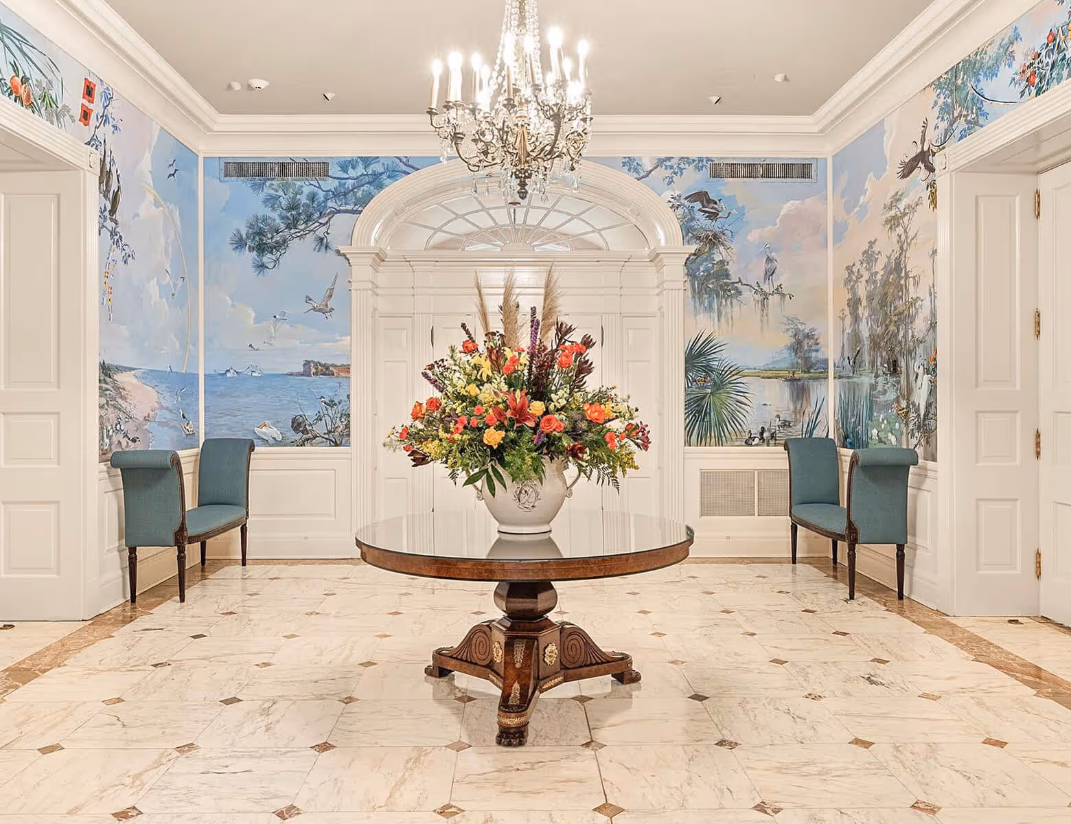 Elegant room with a round wooden table holding a large flower arrangement, chandeliers, blue-green benches, and mural walls depicting nature scenes.