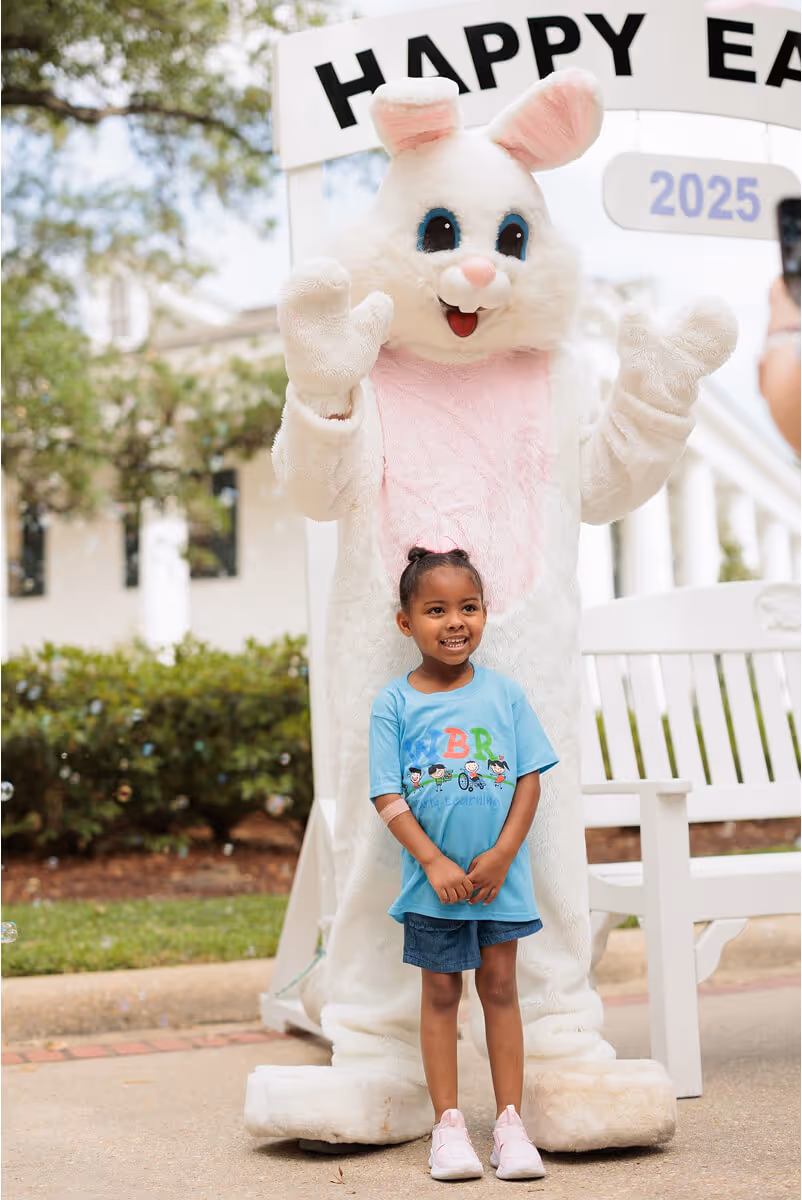 Smiling young girl standing in front of a person dressed as a white Easter bunny with pink ears and a pink chest.
