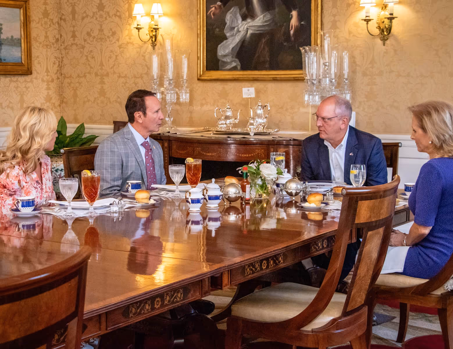 Four people seated at a polished wooden dining table in an elegant room, engaged in conversation with drinks and tableware set before them.