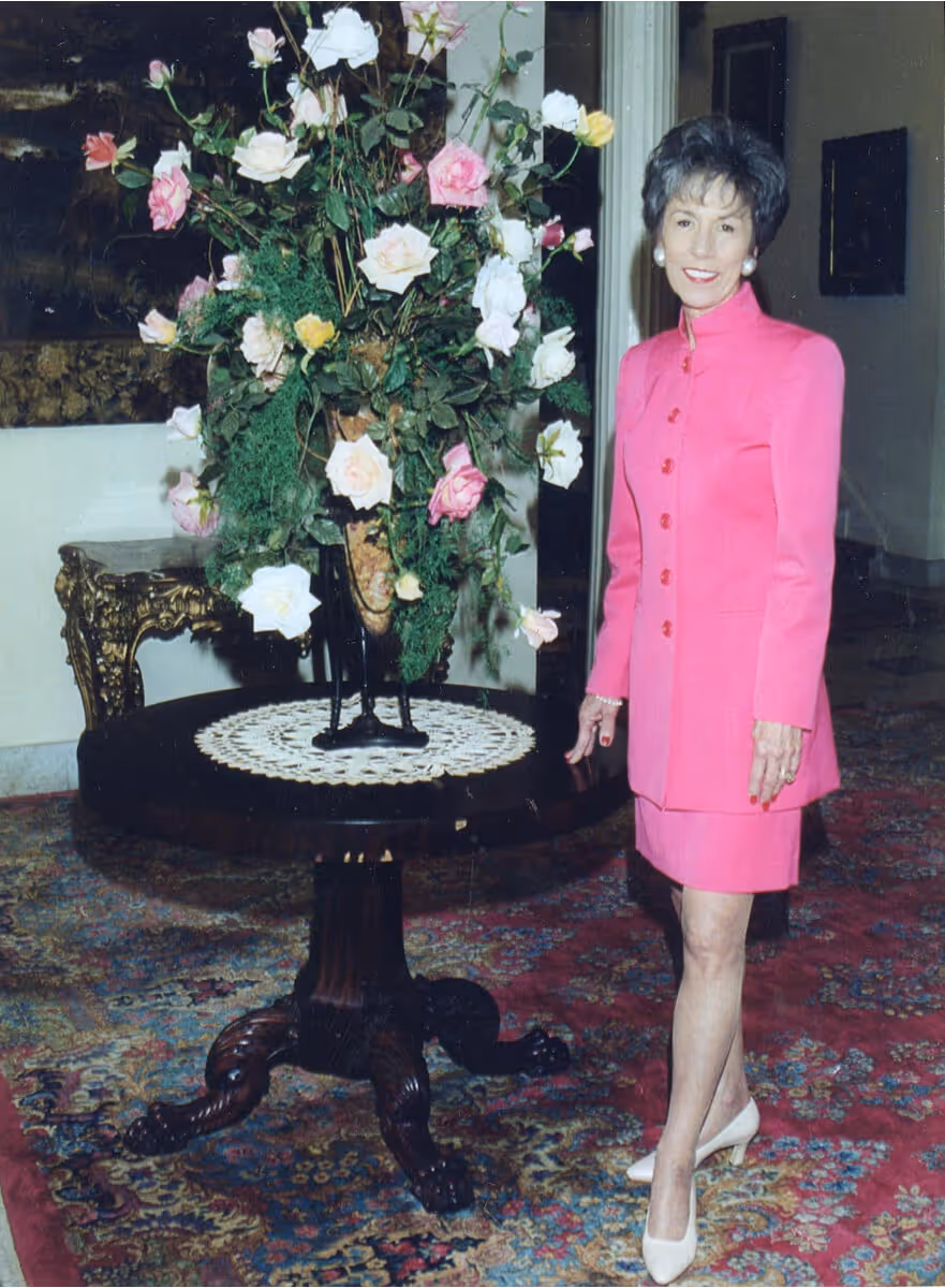 Woman in a bright pink dress standing next to a round wooden table with a large floral arrangement of white, pink, and yellow roses.