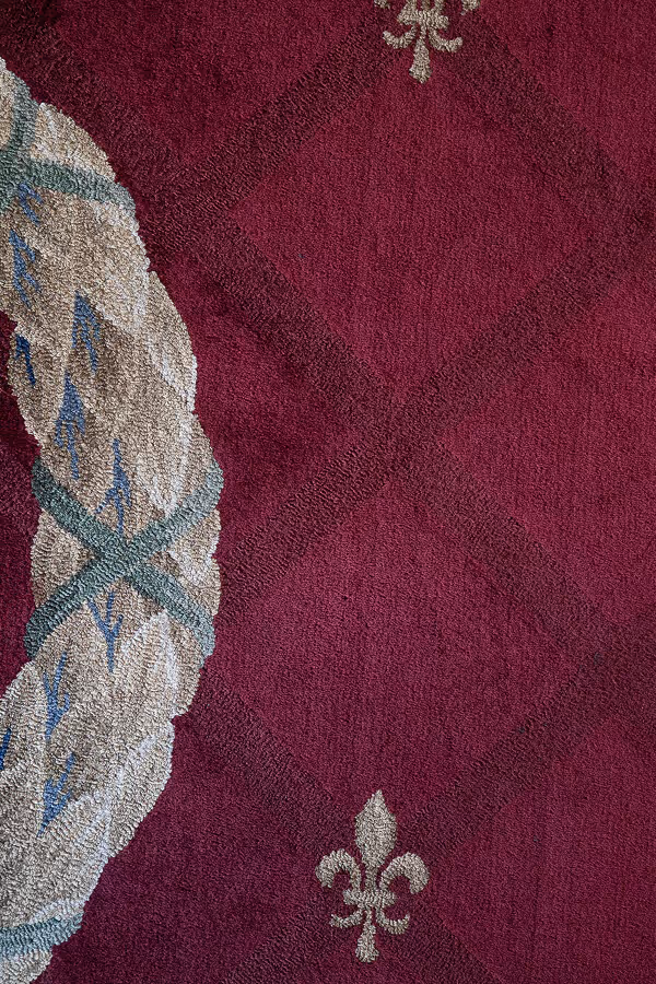 Close-up of a red carpet with diamond patterns, a partial beige and green wreath, and fleur-de-lis motifs.