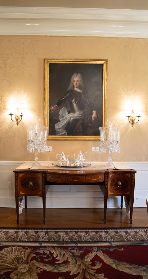 Antique wooden sideboard with silver tea set and crystal candelabras beneath a framed portrait of a man in historic attire, flanked by two wall sconces.