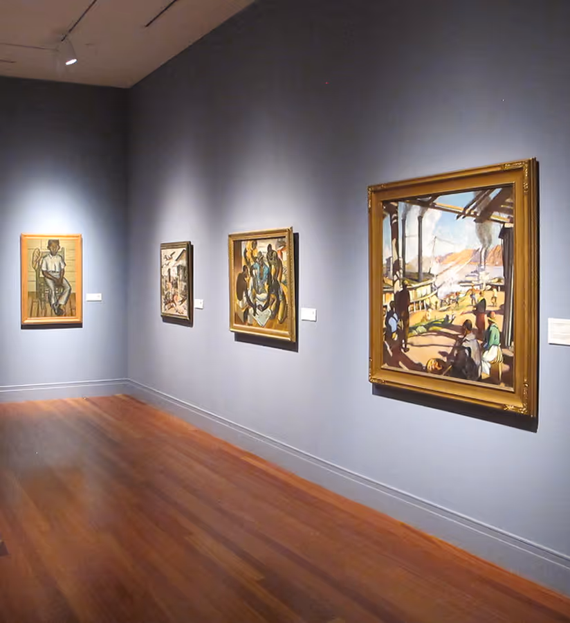 Interior of an art gallery with four framed paintings hanging on a gray wall above wooden flooring.