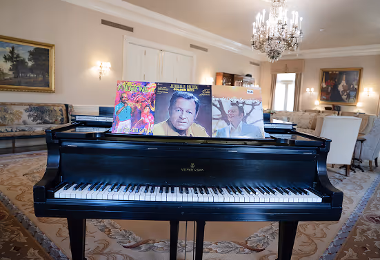 Black grand piano with vinyl records displayed on its music stand in a spacious, elegant living room with chandelier and classic furniture.