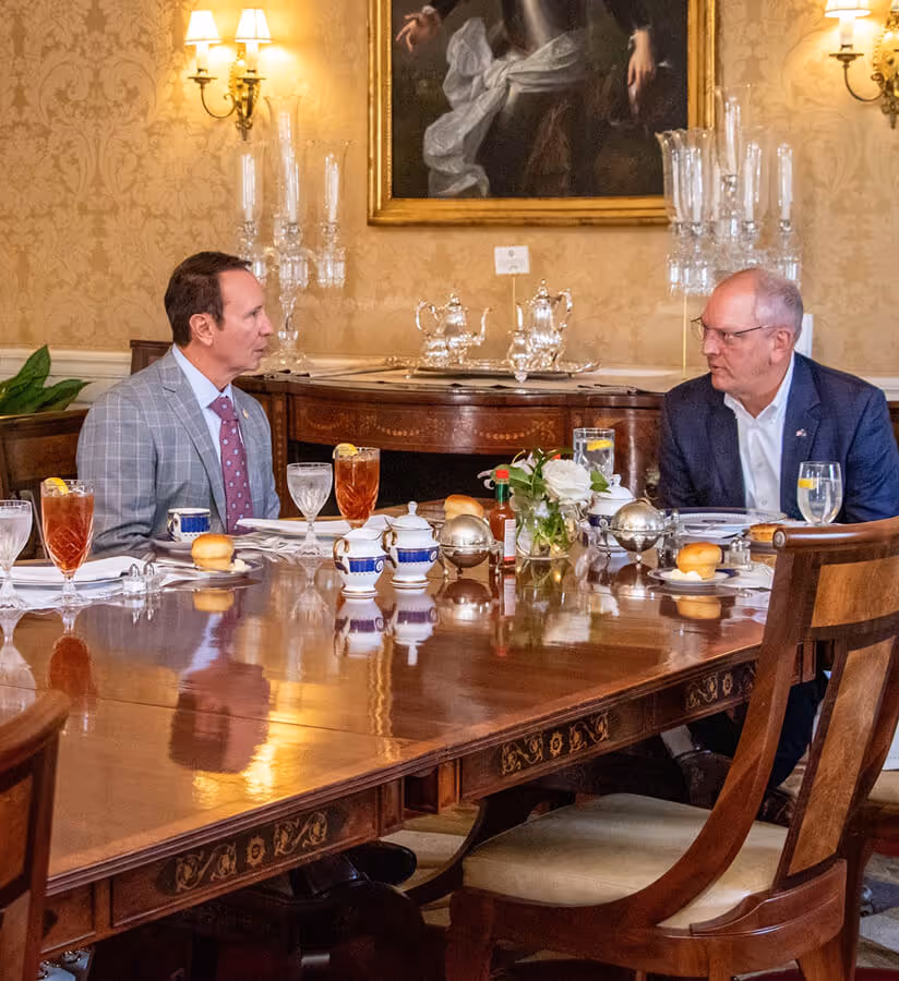 Two men in suits sitting and conversing at a polished wooden dining table set with beverages and tableware in an elegant room with candelabras and a framed portrait.