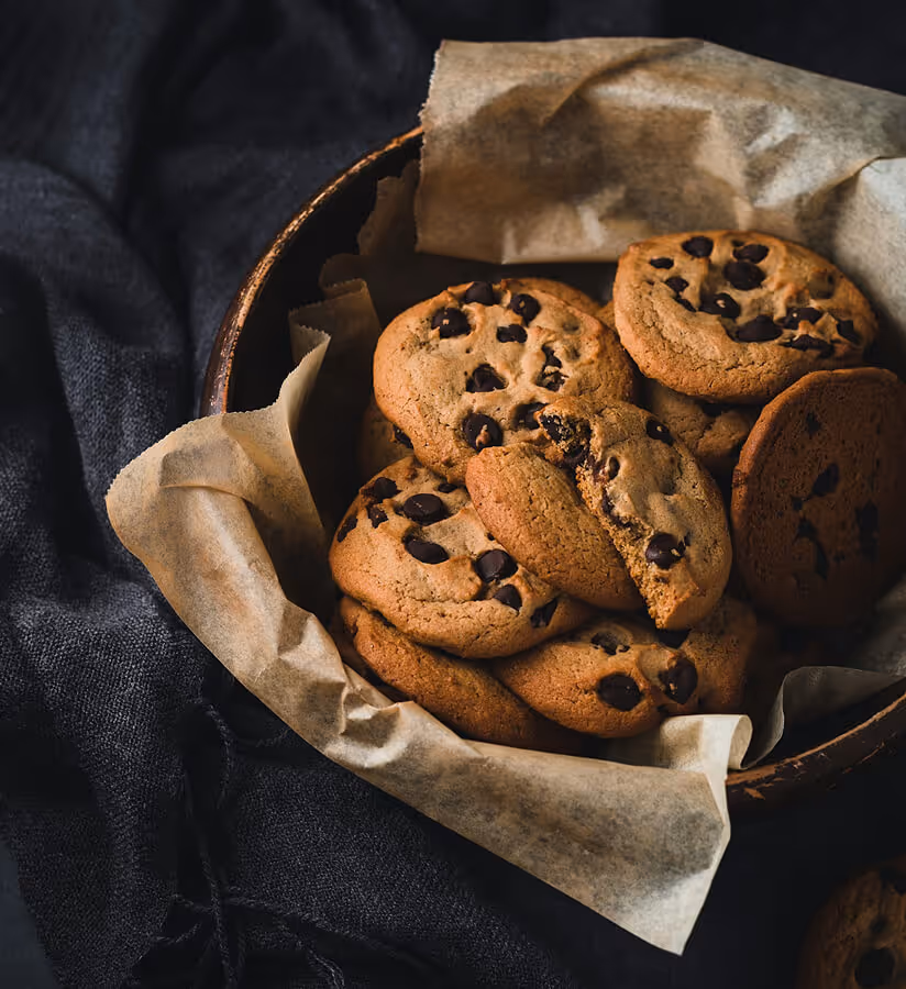 Bowl lined with parchment paper filled with chocolate chip cookies on a dark fabric background.