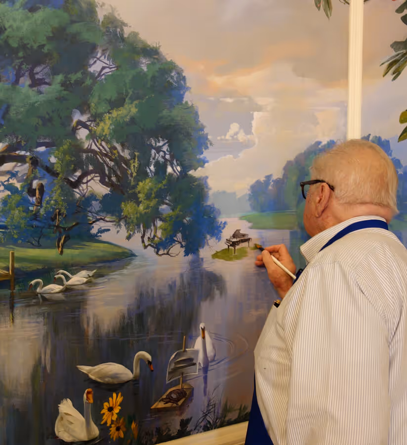 Elderly man with glasses painting a detailed mural of a serene lakeside scene with swans, trees, and a grand piano.