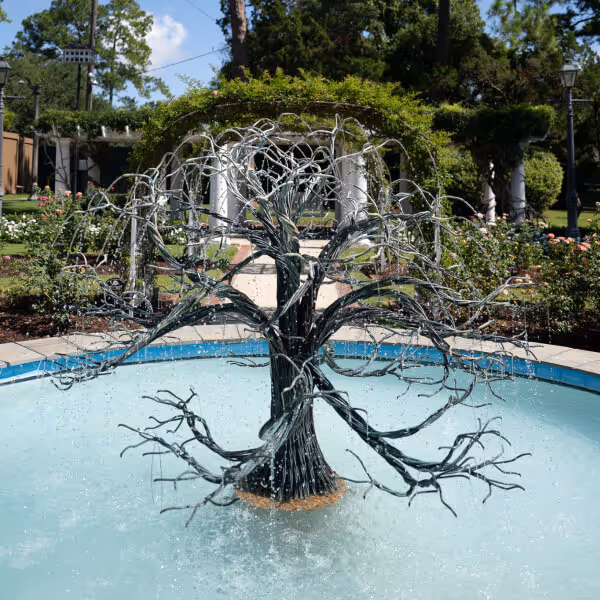 Metal tree-shaped fountain with water dripping from its branches in an outdoor garden pool.