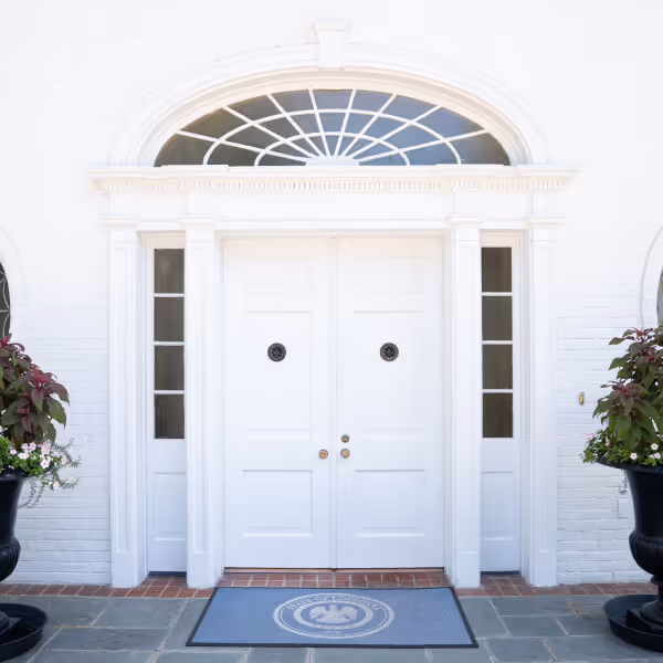 White double front doors with circular windows above and sidelights, flanked by large black planters with green and purple plants.