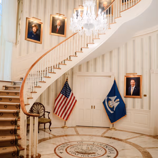 Curved staircase with a chandelier, portraits on the walls, and American and Louisiana state flags flanking double doors in a room with a circular floor seal.