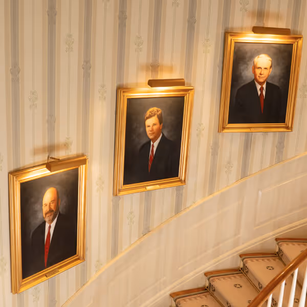 Three framed portraits of men in suits with red ties hanging on a patterned wall above a curved staircase.