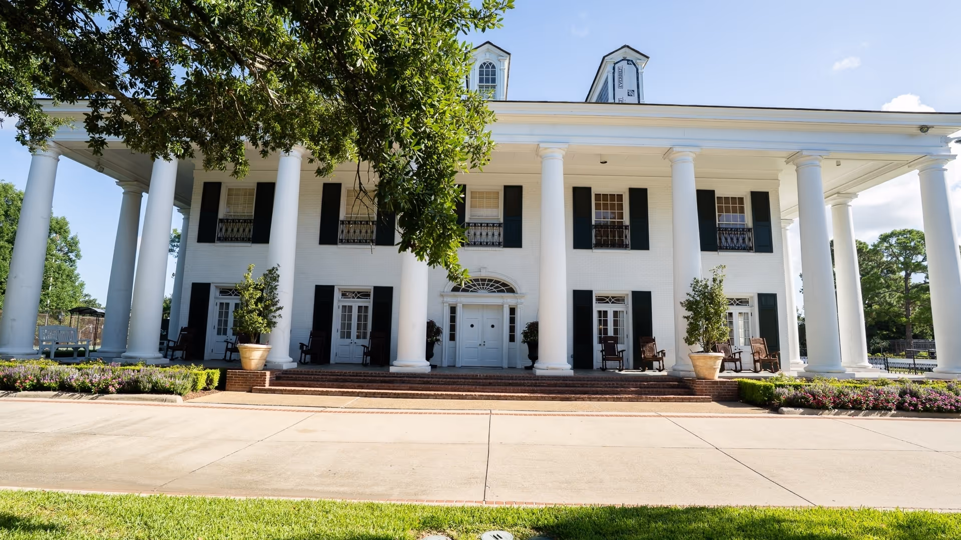 Large white colonial-style house with tall white columns, black shutters, and a covered porch with chairs and potted plants.