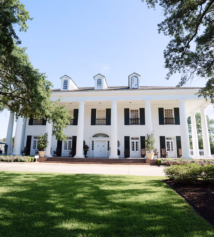 Large white mansion with tall columns, black shutters, and dormer windows set against a clear sky with green lawn and trees in front.