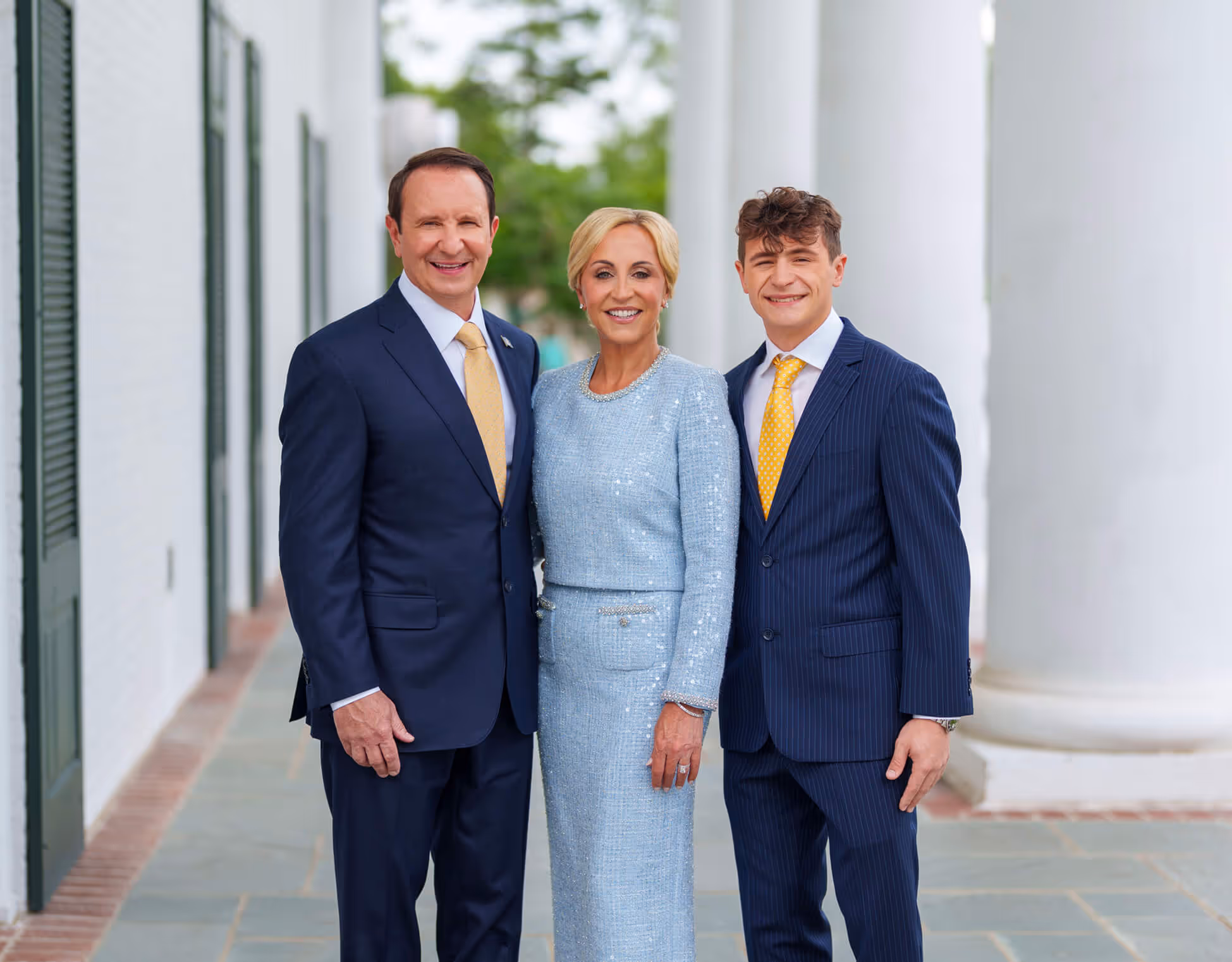 A man, woman, and young man dressed formally standing together outdoors in front of white pillars and smiling at the camera.